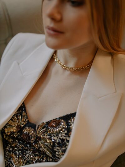 A stylish woman wearing a gold necklace and embellished top with a white blazer indoors.